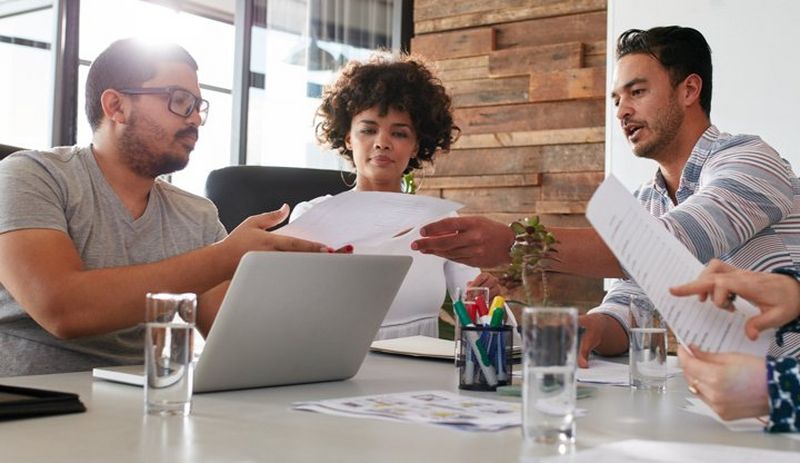 Two men and a woman pass papers over a large white desk, with open laptop, pens in a holder and glasses of water.